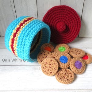 May include: A crocheted toy cookie jar with a lid and several crocheted cookies. The jar is light blue with red and yellow stripes. The lid is red. The cookies are brown with colorful centers.