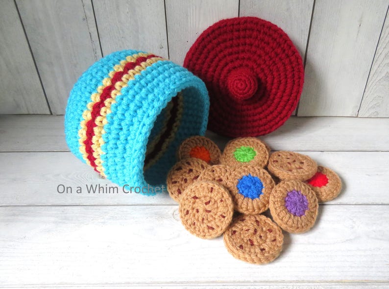 May include: A crocheted toy cookie jar with a lid and several crocheted cookies. The jar is light blue with red and yellow stripes. The lid is red. The cookies are brown with colorful centers.