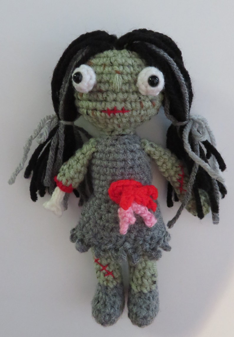 crochet girl zombie with green skin, grey dress and black and grey hair. She has bulging eyes, red scar for mouth, a severed arm with a crochet bone sticking out. She has red and pink intestines coming out of her belly, a red scar one one arm and leg