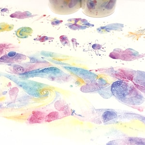 May include: A roll of washi tape with a watercolor galaxy design. The tape features a swirling pattern of pink, blue, and yellow colors with white stars and planets.