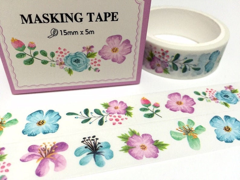May include: A roll of white washi tape with a floral design featuring pink, blue, and green watercolour flowers. The tape is 15mm wide and 5 metres long. The tape is packaged in a pink box with the text "MASKING TAPE" and "15mm x 5m".