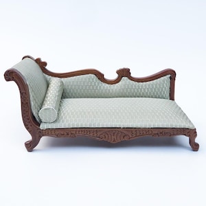 Puede incluir: Chaise longue en miniatura con un cojín estampado verde claro y un cojín cilíndrico a juego. El marco de madera es de color marrón oscuro con tallas ornamentadas. Este pequeño sofá es una pieza decorativa.
