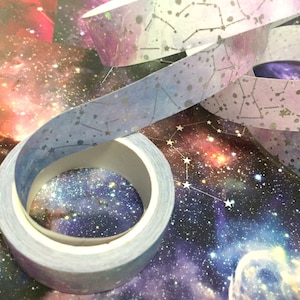 Può includere: Un rotolo di nastro washi con una stampa galattica e stelle argentate scintillanti. Il nastro è blu, viola e bianco con un effetto acquerello.