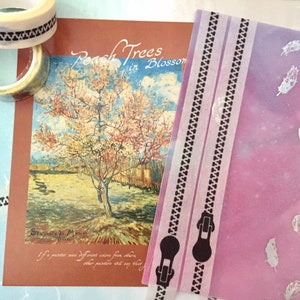 May include: A pink and white patterned sheet of paper with a black zipper design and white feathers printed on it. The paper is folded in half and has a white border. There are two rolls of washi tape in the background.