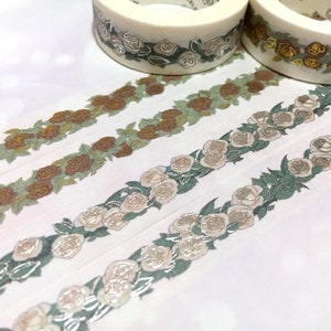 以下が含まれることがあります： 花柄の和紙テープが4ロール。テープには、緑、茶色、金色のバラと葉の繰り返し模様が施されています。テープは白い背景の上にあります。