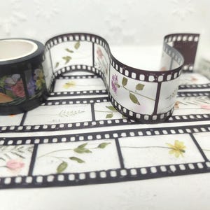Może przedstawiać: Dekoracyjna taśma washi z kwiatowym wzorem taśmy filmowej. Czarna rolka taśmy przedstawia kolorowe kwiaty, a wz&oacute;r taśmy filmowej ma białe tło z kwiatowymi akcentami. Widoczna jest r&oacute;wnież rolka przezroczystej taśmy.