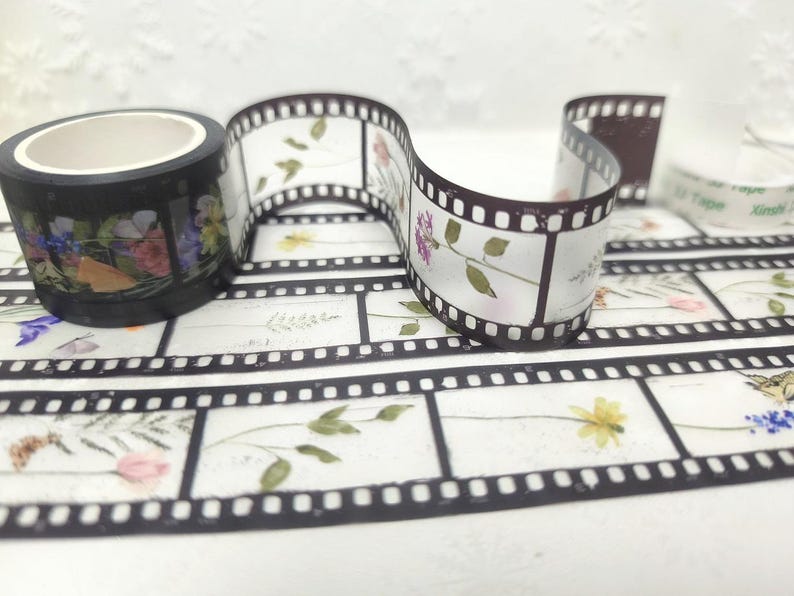 Może przedstawiać: Dekoracyjna taśma washi z kwiatowym wzorem taśmy filmowej. Czarna rolka taśmy przedstawia kolorowe kwiaty, a wz&oacute;r taśmy filmowej ma białe tło z kwiatowymi akcentami. Widoczna jest r&oacute;wnież rolka przezroczystej taśmy.