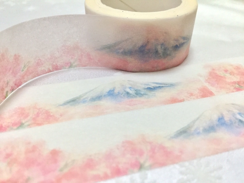K&ouml;nnte beinhalten: Eine Rolle Washi-Tape mit einem Aquarell-Design aus einer Bergkette und rosa Blumen. Das Band ist transparent und hat einen wei&szlig;en Hintergrund.