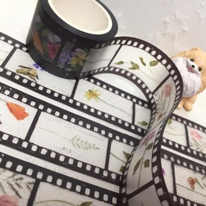 Może przedstawiać: Rolka taśmy washi z czarnym tłem i wzorami kwiatowymi i motylami. Widoczny jest r&oacute;wnież pasek taśmy washi w stylu filmowym z ilustracjami kwiat&oacute;w i owad&oacute;w. Obecna jest r&oacute;wnież mała figurka psa.