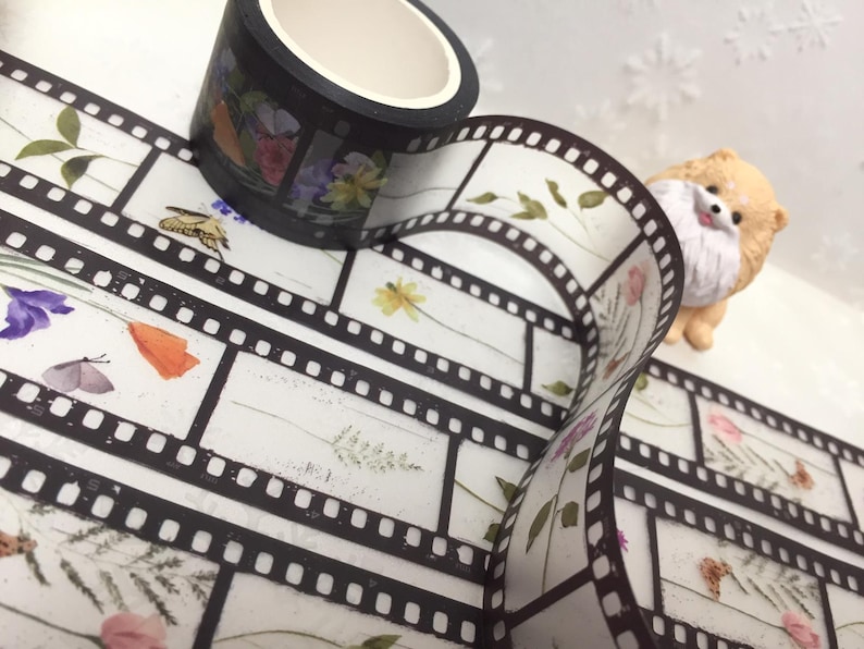 Może przedstawiać: Rolka taśmy washi z czarnym tłem i wzorami kwiatowymi i motylami. Widoczny jest r&oacute;wnież pasek taśmy washi w stylu filmowym z ilustracjami kwiat&oacute;w i owad&oacute;w. Obecna jest r&oacute;wnież mała figurka psa.