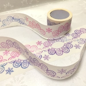 May include: A roll of white washi tape with a repeating pattern of purple and pink lace designs and snowflakes. The tape is on a white surface.