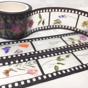 Może przedstawiać: Dekoracyjna taśma washi z motywem taśmy filmowej z kwiatowymi i motylkowymi akcentami. Czarna rolka taśmy ma biały rdzeń. Wz&oacute;r taśmy filmowej ma przezroczyste tło z kolorowymi prasowanymi kwiatami i owadami.