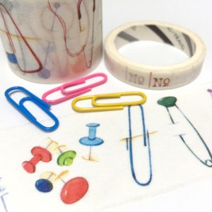 May include: Two rolls of washi tape with different patterns. One roll has a white background with colorful geometric shapes and the other has a white background with colorful pins, safety pins, and paper clips. The tape is labeled "NO NO".
