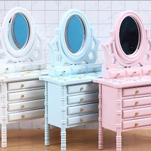 May include: Three miniature wooden vanity tables in white, light blue, and pink. Each has an oval mirror, three small drawers, and four larger drawers with gold-colored knobs. The tables are decorated with floral patterns.