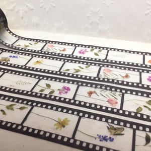Może przedstawiać: Dekoracyjna taśma washi z motywem taśmy filmowej ozdobiona motywami kwiatowymi i motylami. Czarna taśma ma białe tło, a wz&oacute;r taśmy filmowej zawiera r&oacute;żne kwiaty i owady.