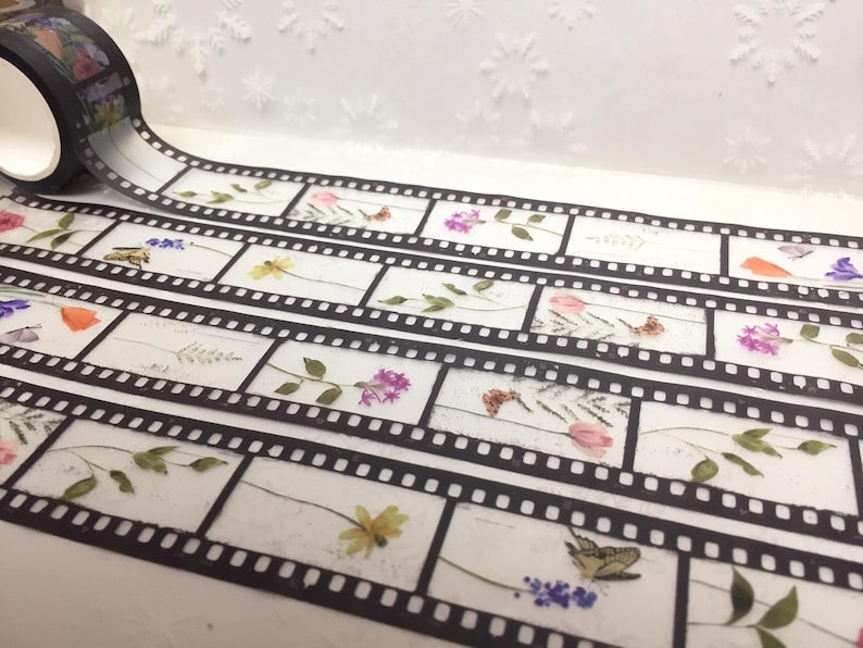 Może przedstawiać: Dekoracyjna taśma washi z motywem taśmy filmowej ozdobiona motywami kwiatowymi i motylami. Czarna taśma ma białe tło, a wz&oacute;r taśmy filmowej zawiera r&oacute;żne kwiaty i owady.