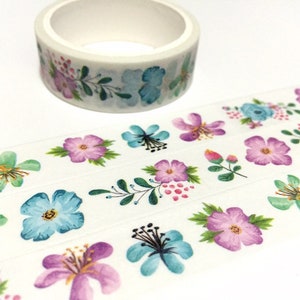 May include: A roll of white washi tape with a floral pattern. The tape features blue, pink, and purple flowers with green leaves on a white background.