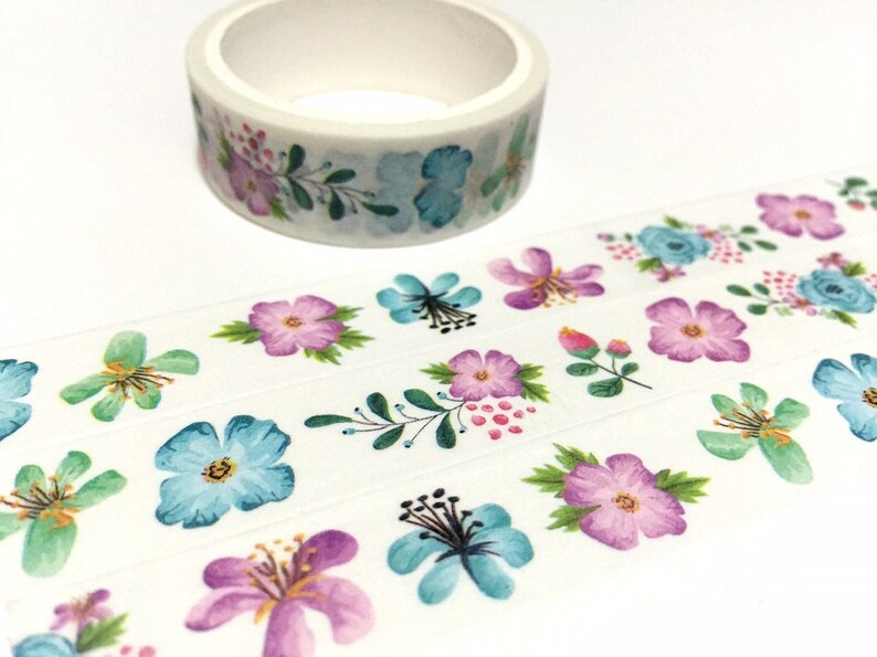 May include: A roll of white washi tape with a floral pattern. The tape features blue, pink, and purple flowers with green leaves on a white background.