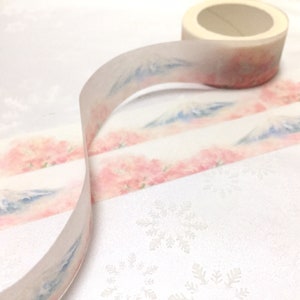 K&ouml;nnte beinhalten: Eine Rolle Washi-Tape mit einem zarten Aquarell-Design mit rosa Bl&uuml;ten und einer blauen Bergkette. Das Band ist auf einer wei&szlig;en Oberfl&auml;che mit einem Schneeflockenmuster abgewickelt.