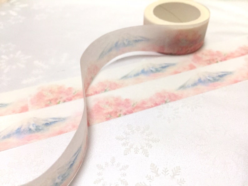 K&ouml;nnte beinhalten: Eine Rolle Washi-Tape mit einem zarten Aquarell-Design mit rosa Bl&uuml;ten und einer blauen Bergkette. Das Band ist auf einer wei&szlig;en Oberfl&auml;che mit einem Schneeflockenmuster abgewickelt.