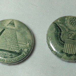 Puede incluir: Dos botones redondos verdes y blancos con imágenes del reverso de un billete de dólar estadounidense. Un botón presenta la pirámide con el ojo que todo lo ve y el texto "ANNUIT COEPTIS" y "NOVUS ORDO SECLORUM". El otro botón presenta el águila con el texto "E PLURIBUS UNUM".
