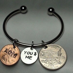 May include: A silver bangle bracelet with three charms: a copper penny with the word "LOVE" engraved on it, a silver charm with "YOU & ME" engraved on it, and a silver quarter with the state of Arizona on it.