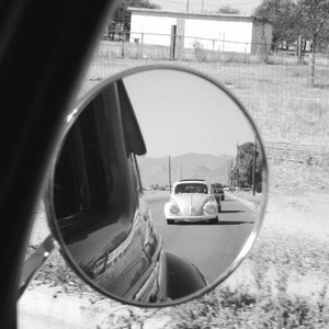 Puede incluir: Una foto en blanco y negro de un espejo retrovisor que refleja un Volkswagen Beetle blanco conduciendo por una carretera. El coche está en la distancia y el espejo refleja la vista lateral del coche.
