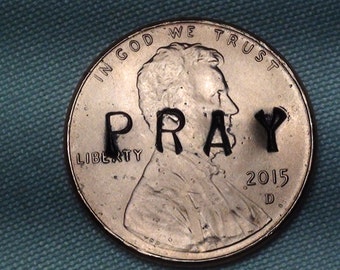 Puede incluir: Primer plano de un centavo estadounidense con las palabras "IN GOD WE TRUST" y "LIBERTY" inscritas en él. El año "2015" también es visible. La palabra "PRAY" está escrita con marcador negro en la moneda.