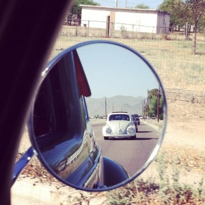Puede incluir: Un espejo retrovisor redondo con marco negro que refleja un Volkswagen Beetle blanco conduciendo por una carretera con otros coches.