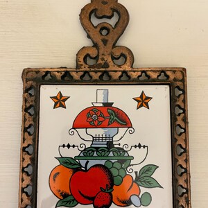 May include: A decorative trivet with a copper-colored metal frame and handle. The square tile insert features a colorful illustration of a lamp, fruit, and stars. The trivet measures approximately 7 inches square.