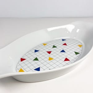 May include: White oval serving dish with a grid pattern and colourful triangles. The dish has a smooth, glossy finish and a slightly raised edge. The triangles are red, yellow, green, and blue.
