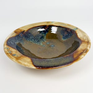 May include: A handmade ceramic bowl with a wide, shallow design. The bowl features a glossy, dark brown interior and a rim with a blend of tan, brown, and blue-green hues. The bowl's surface has a textured appearance.