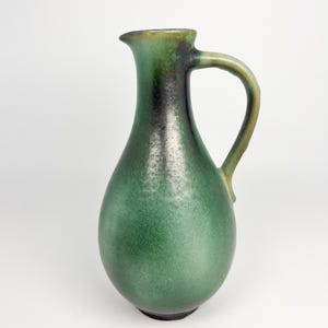 Puede incluir: Jarra de cerámica verde con asa. La jarra tiene un cuerpo bulboso y un cuello estrecho con pico. El asa es curva y está unida al lateral. La jarra está esmaltada con un acabado verde moteado.