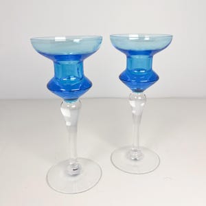 May include: Two glass candle holders with a distinctive design. They feature a clear base and stem, a bright blue body, and a light blue top. The design is modern and elegant, suitable for home decor.