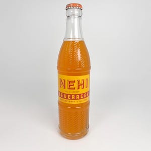 Puede incluir: Una botella de vidrio vintage de Nehi Beverages, llena de un líquido de color naranja. La botella tiene un diseño texturizado y una etiqueta amarilla con letras rojas. La tapa de la botella es naranja. La etiqueta dice "NEHI Beverages, Contenido neto 355 ml."