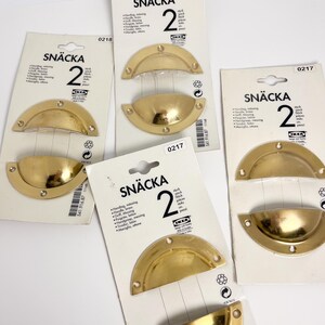 May include: Several packages of IKEA SNÄCKA brass-colored drawer pulls. Each package contains two pulls. The pulls are a half-moon shape with pre-drilled holes for mounting. The packaging includes the IKEA logo and product information.
