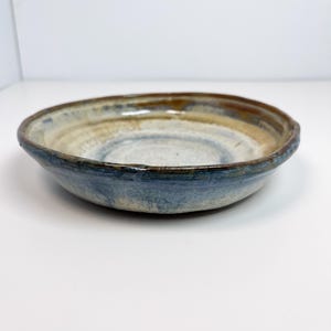 May include: A handcrafted ceramic bowl with a wide, shallow form. The bowl displays a mix of colours, including blue, brown, and cream, with a textured finish. The rim has a darker brown tone, and the bowl is suitable for serving or display.