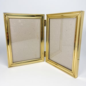 May include: A double-hinged picture frame with a polished gold-tone metal finish. The frame consists of two rectangular sections connected by two vertical hinges, each containing a light beige mat board insert within the metallic border.
