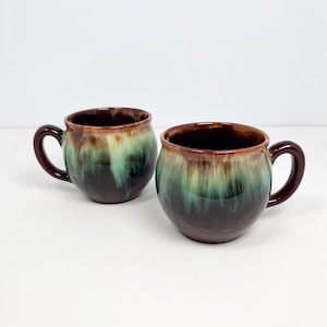 May include: Two ceramic mugs with a rounded shape and dark brown handles. The mugs have a glossy finish with a colour gradient of brown, green, and tan. The mugs are designed for beverages.