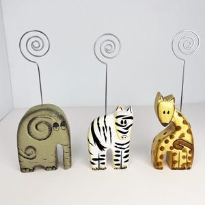 May include: Three wooden animal photo holders with metal wire loops. The holders are painted to look like an elephant, zebra, and giraffe. The elephant is green, the zebra is black and white, and the giraffe is yellow with brown spots.