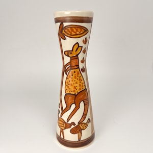 May include: A tall, cylindrical ceramic vase with a cream-colored base and brown painted details. The design features a central kangaroo figure surrounded by stylized plant motifs and smaller animal shapes in shades of brown and orange.