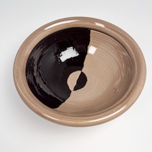 May include: A handmade ceramic bowl with a unique design. The interior is split into two halves: one in a deep black glaze and the other in a light tan. The rim and exterior are a matching tan color. The bowl is round and has a simple, elegant shape.