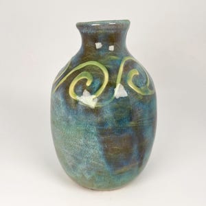 May include: A small ceramic vase with a rounded body and a narrow neck. The surface features a blended glaze of deep blue, teal, and earthy brown tones, accented by a prominent light green swirling spiral pattern and small white flecks.