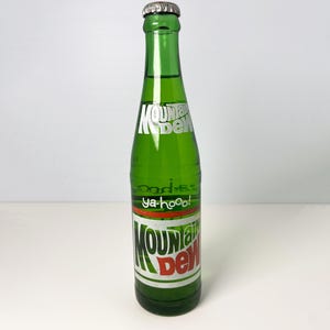 Puede incluir: Una botella de refresco Mountain Dew de vidrio verde con tapa plateada. La botella tiene una etiqueta blanca, roja y verde con el logotipo de Mountain Dew y el texto "ya-hoo!".