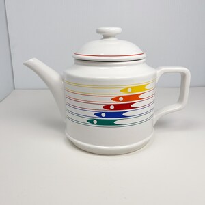 May include: White ceramic teapot with a lid and handle. The teapot is decorated with horizontal bands of color, including red, yellow, blue, and green, with matching dots. The lid has a red trim.