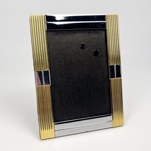 May include: A rectangular picture frame featuring a combination of polished silver-tone metal and vertical ribbed gold-tone metal accents. The frame surrounds a black textured insert with two small circular cutouts near the top center.