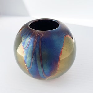 May include: A small, round ceramic vase with a metallic glaze. The glaze has a blue, green, and gold color scheme with a red accent.