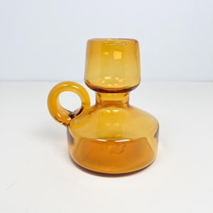 Könnte beinhalten: Bernsteinfarbener Glaskrug mit rundem Boden, zylindrischem Oberteil und kreisförmigem Griff. Der Krug besteht aus transparentem Glas, das Licht durchlässt. Das Design ist schlicht und elegant, geeignet zum Servieren von Getränken oder als Dekoration.