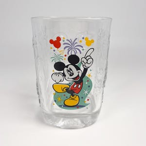 May include: Clear glass tumbler featuring a colorful image of Mickey Mouse in a celebratory pose. The design includes red, yellow, and green accents, with a fireworks and confetti background. The glass has a textured pattern on the sides.