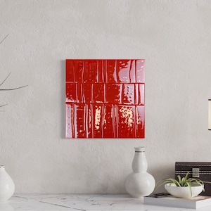 May include: A square, glossy red wall tile installation with a textured surface. The tile is mounted on a light-coloured wall. Decorative items, including vases, a lamp, and a small box, are on a surface below.